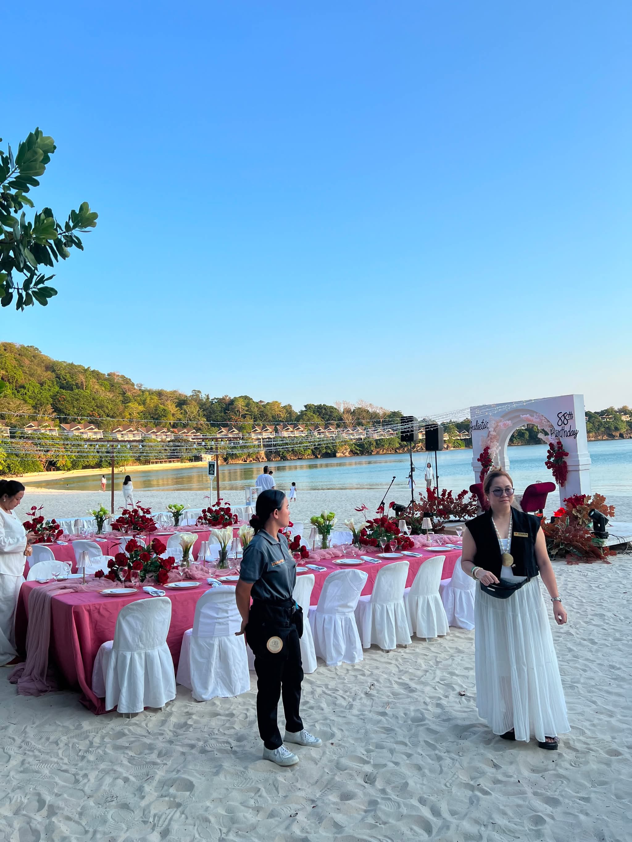 A coordinator standing beside a long beachside table dressed in pink linen and red roses, a floral arch at its head