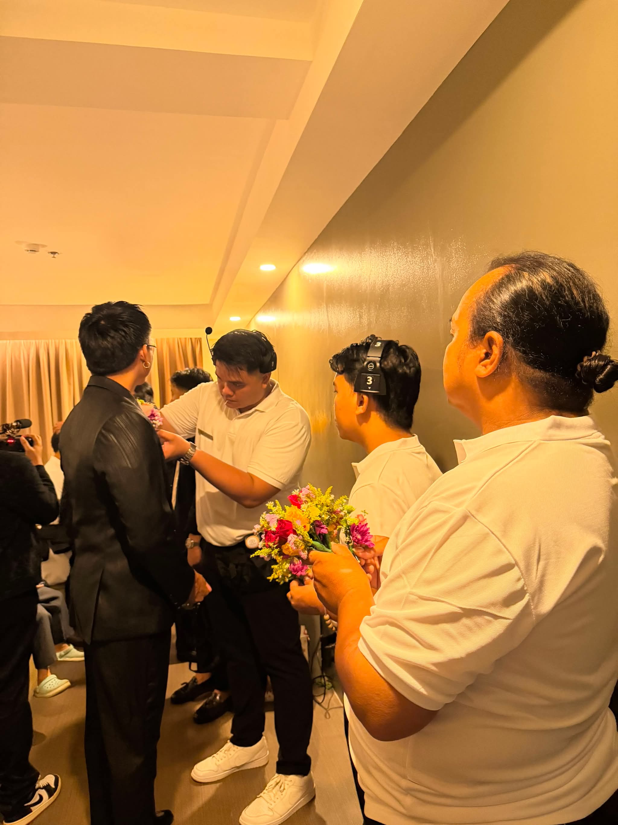 Crew in white polos pinning a boutonniere to the groom's lapel while another holds the flowers