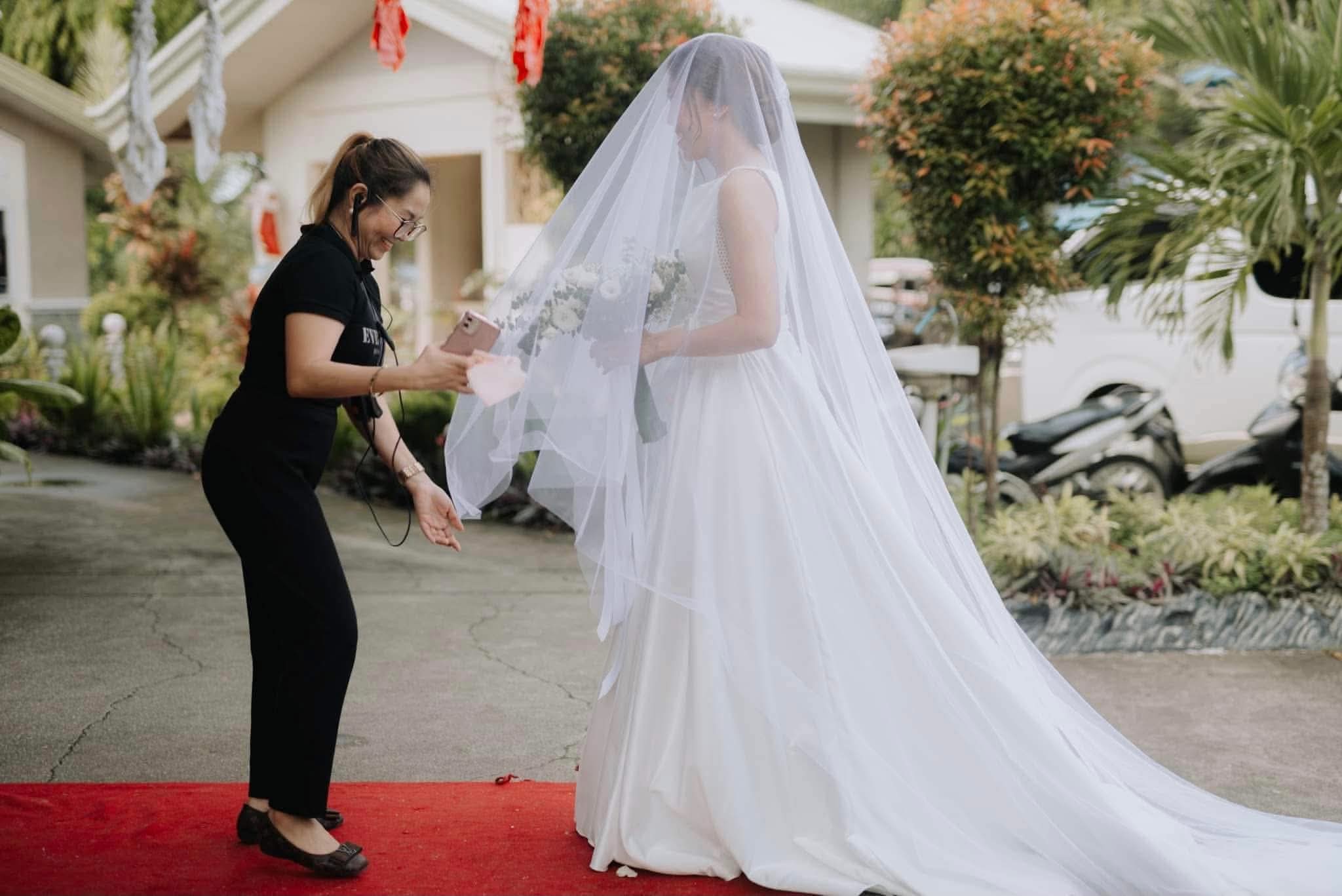 A coordinator in a headset holding a phone out to a veiled bride waiting on a red carpet