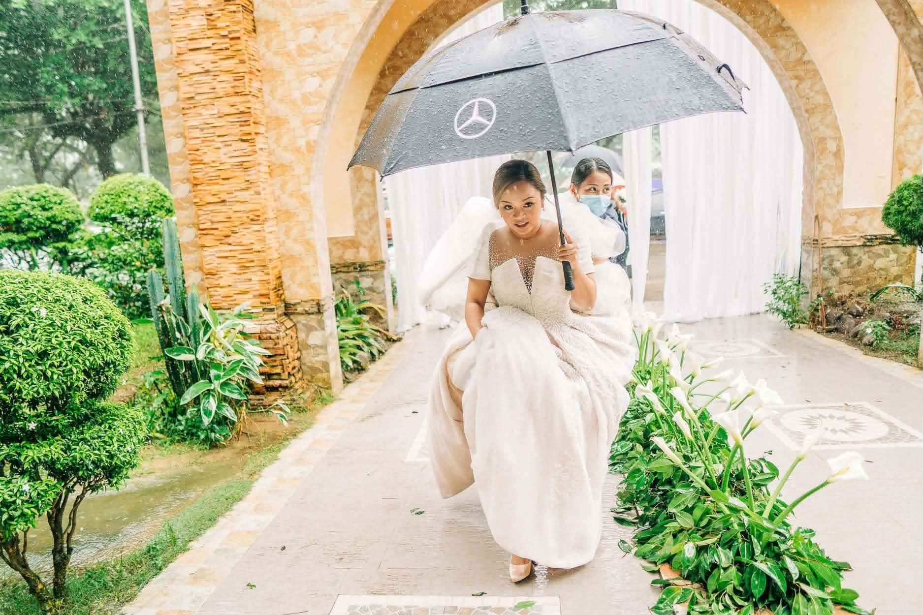 A bride gathering her skirt and hurrying up a garden path in the rain, a coordinator walking behind her holding an umbrella over her
