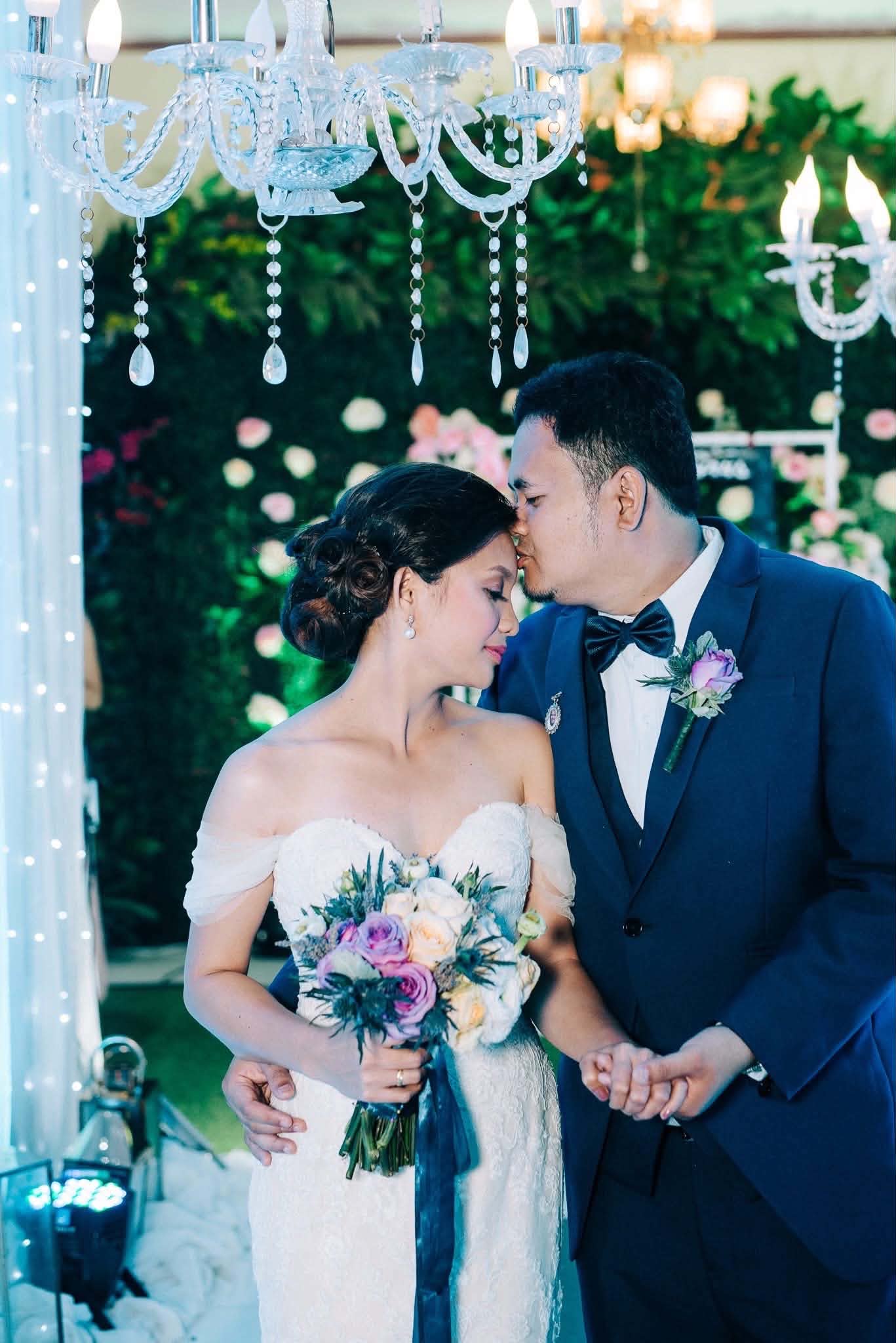 A groom kissing his bride's forehead beneath crystal chandeliers