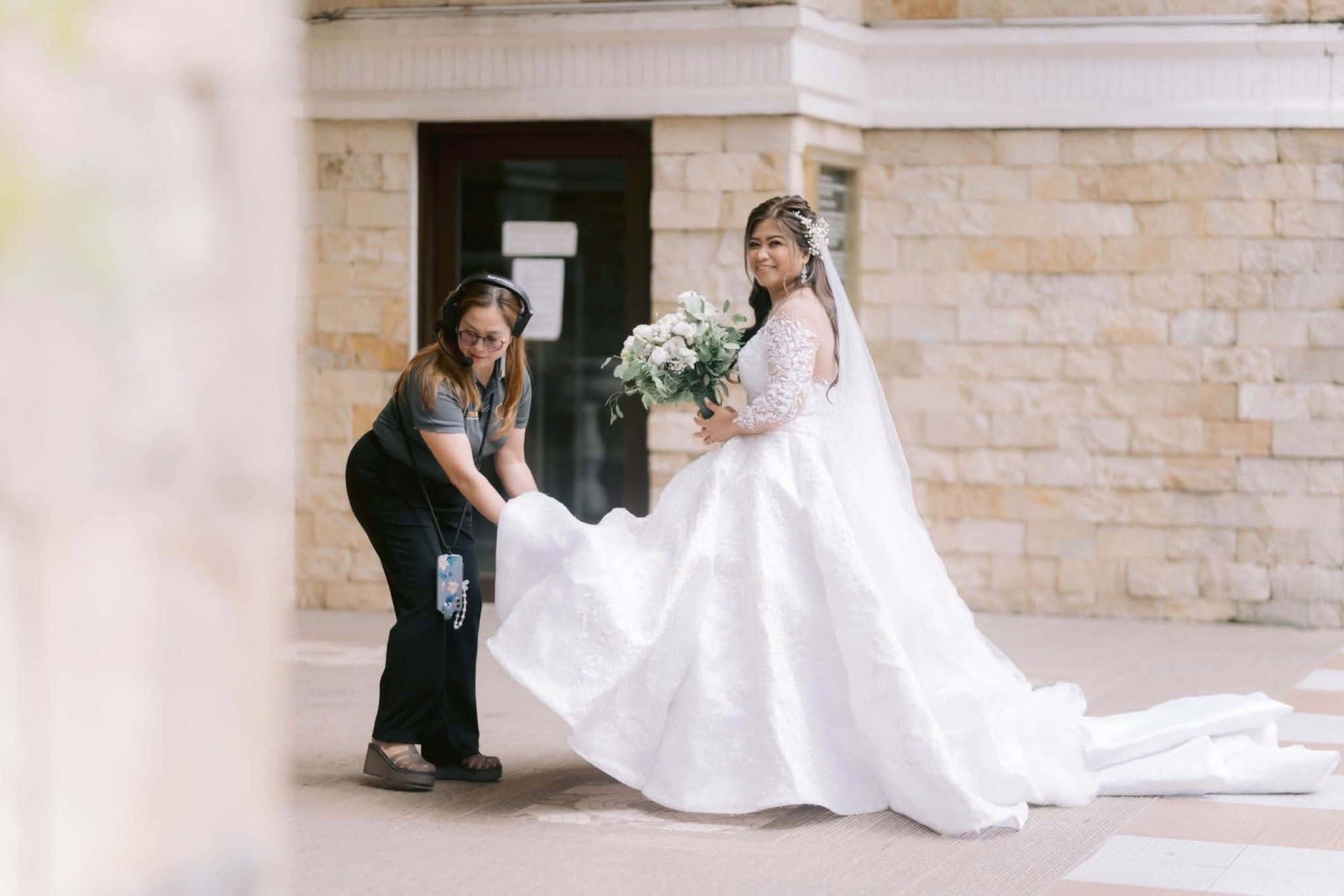 A coordinator in a headset crouching to fan out the bride's train outside a stone church