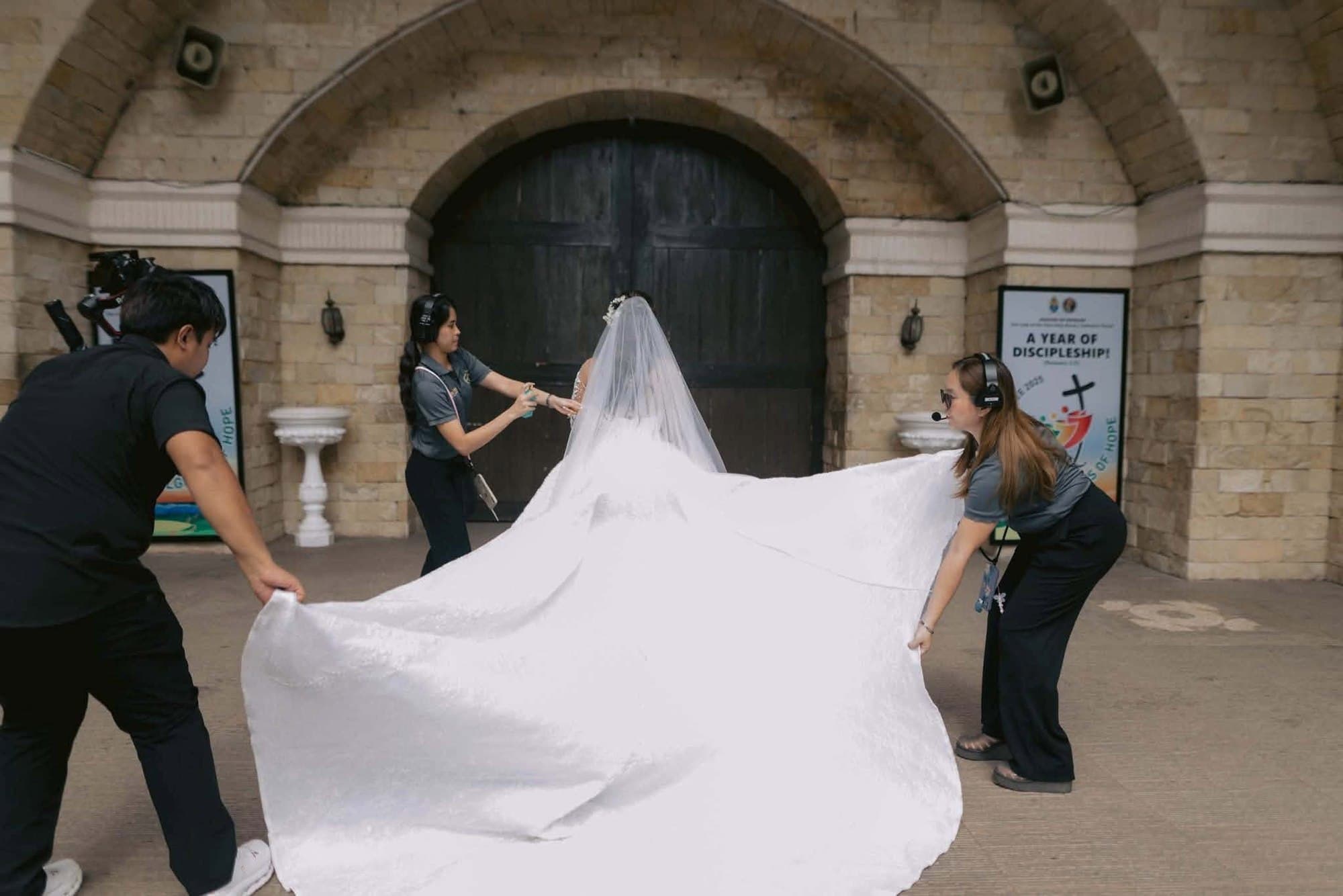 Three crew members spreading a cathedral-length train at the doors of a church while a videographer films