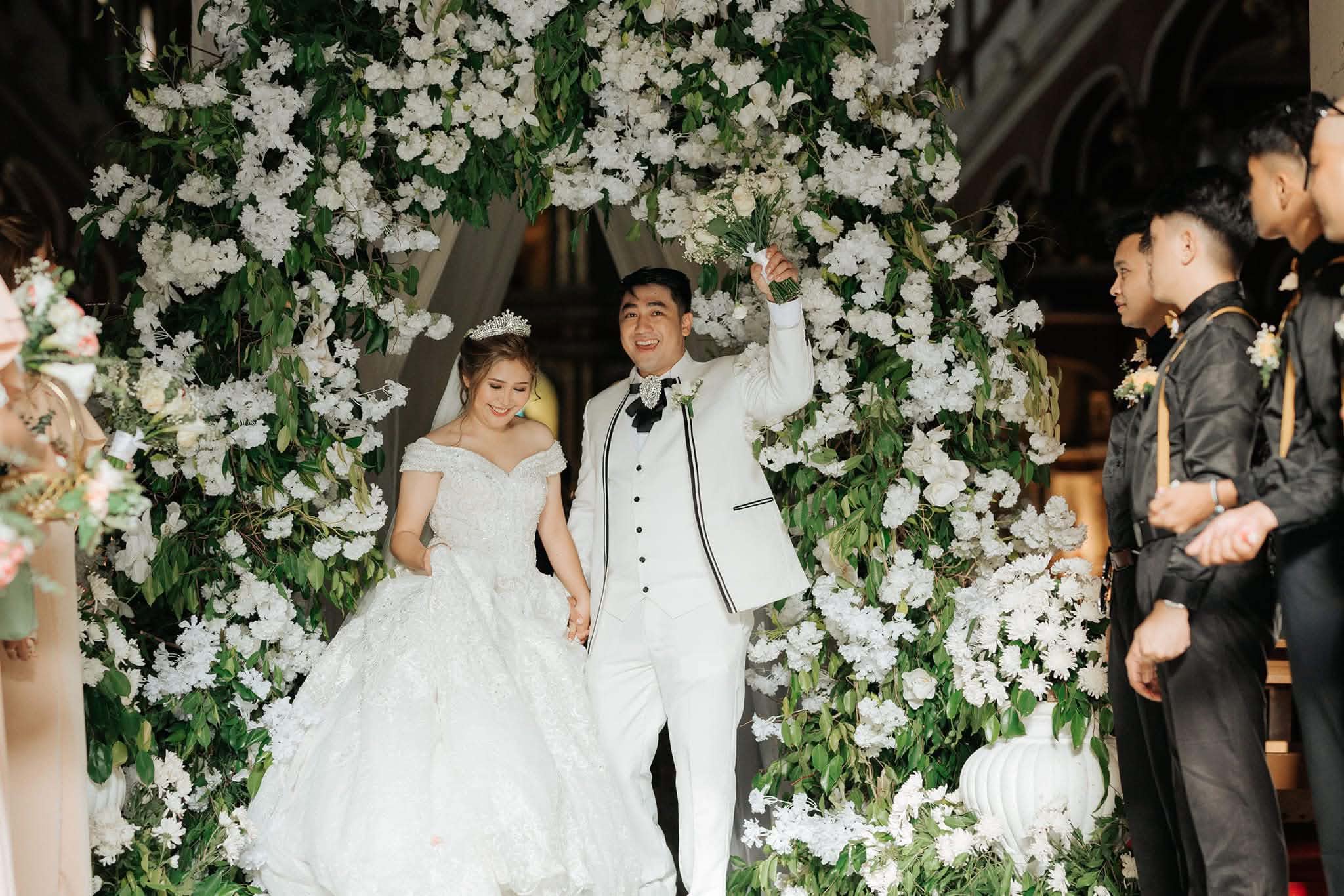 The newlyweds stepping out beneath an arch of white blooms and greenery, the groom holding the bouquet aloft as the entourage looks on