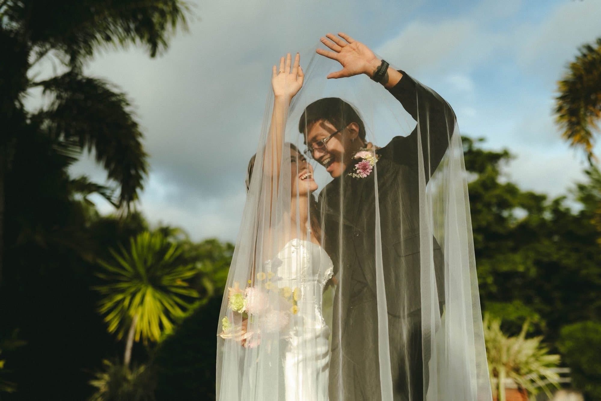 A couple laughing beneath a lifted veil, palms and sky behind them