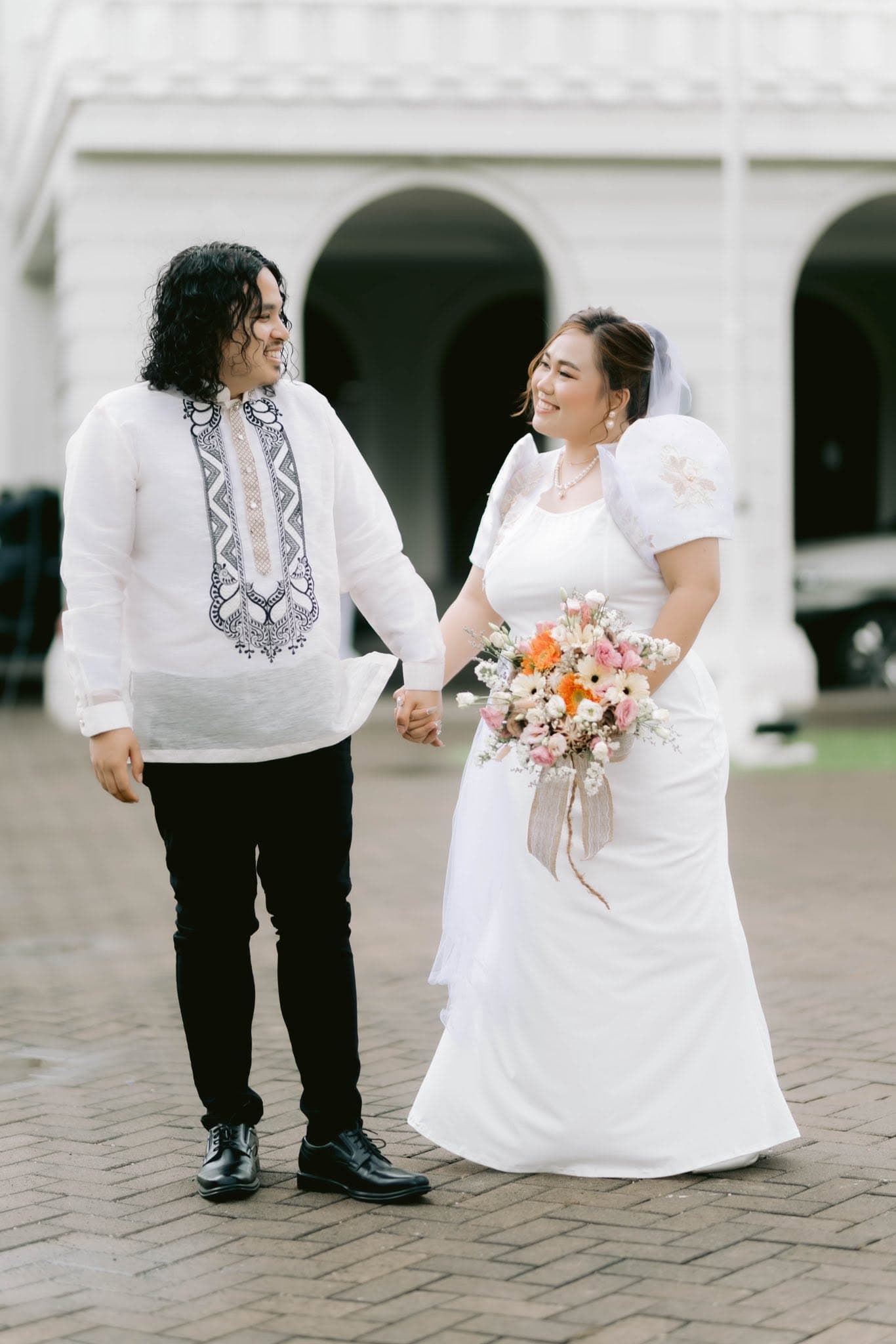 Kyla and Anou hand in hand outside a white colonial hall, he in an embroidered barong and she in a white gown with a bouquet