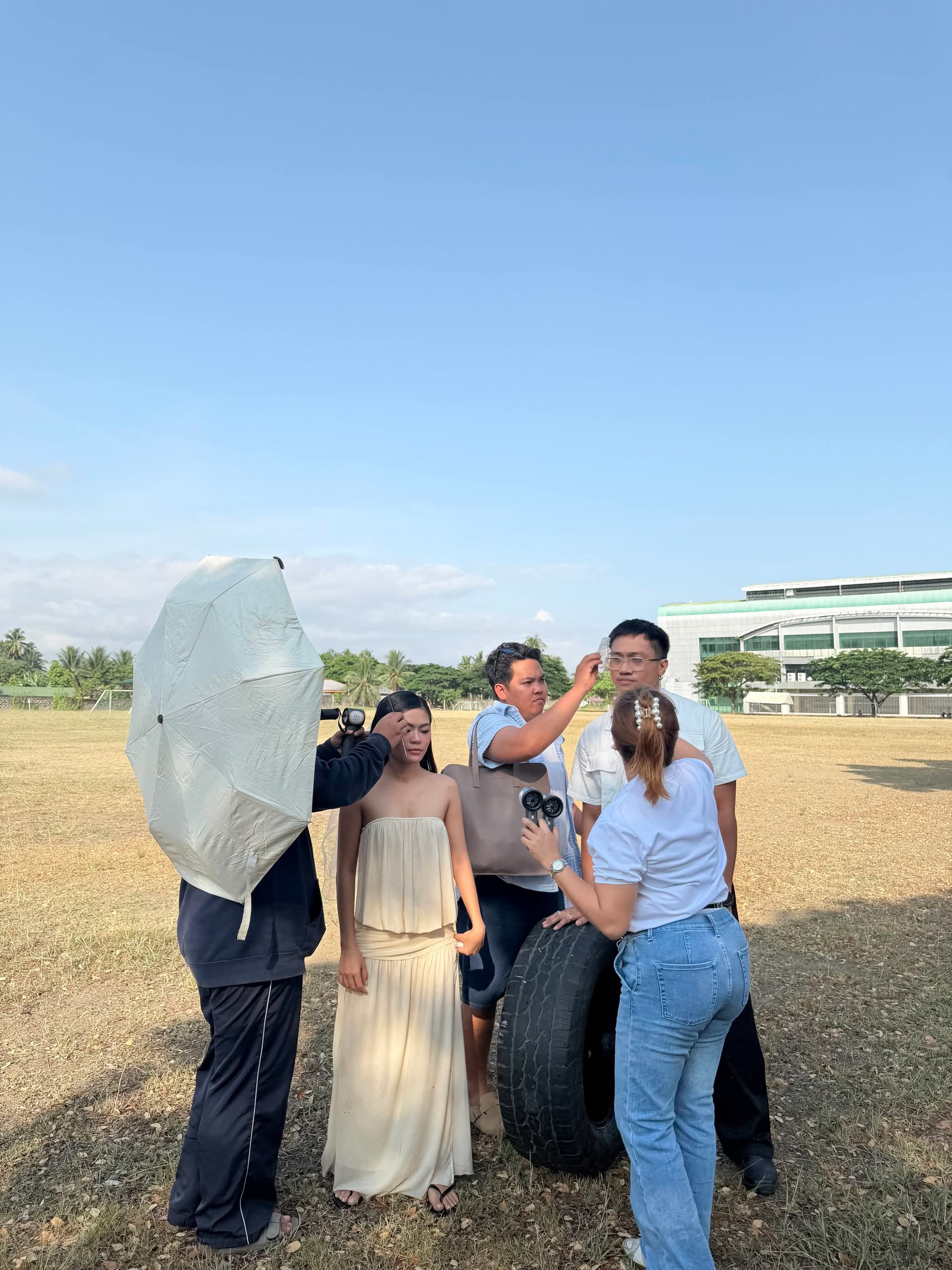 Crew on an open field shading a model with a reflector umbrella, holding a fan to her and touching up her hair