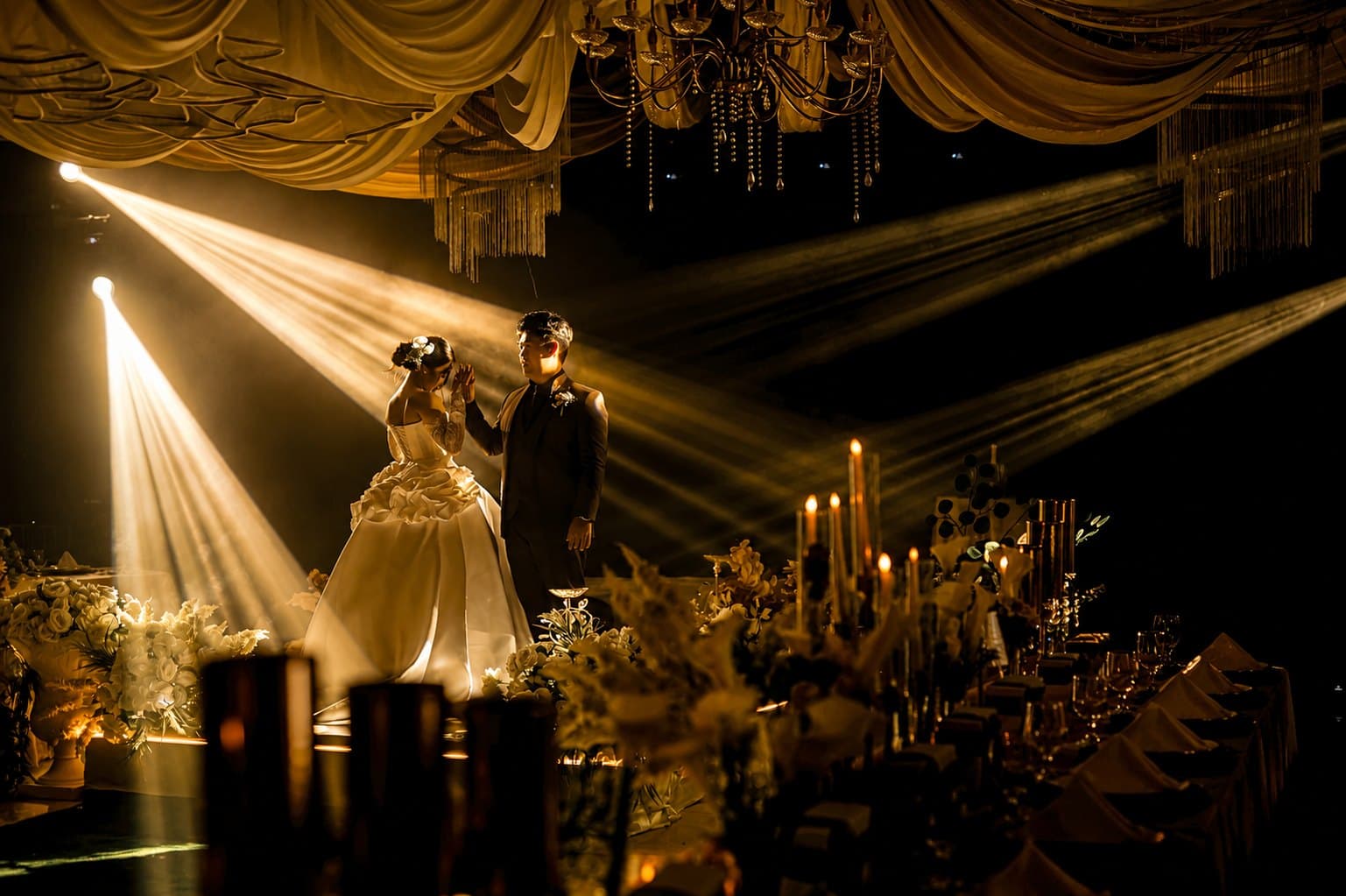 A newlywed couple mid-dance on a candlelit stage under crystal chandeliers, spotlight beams crossing the dark room above a long banquet table