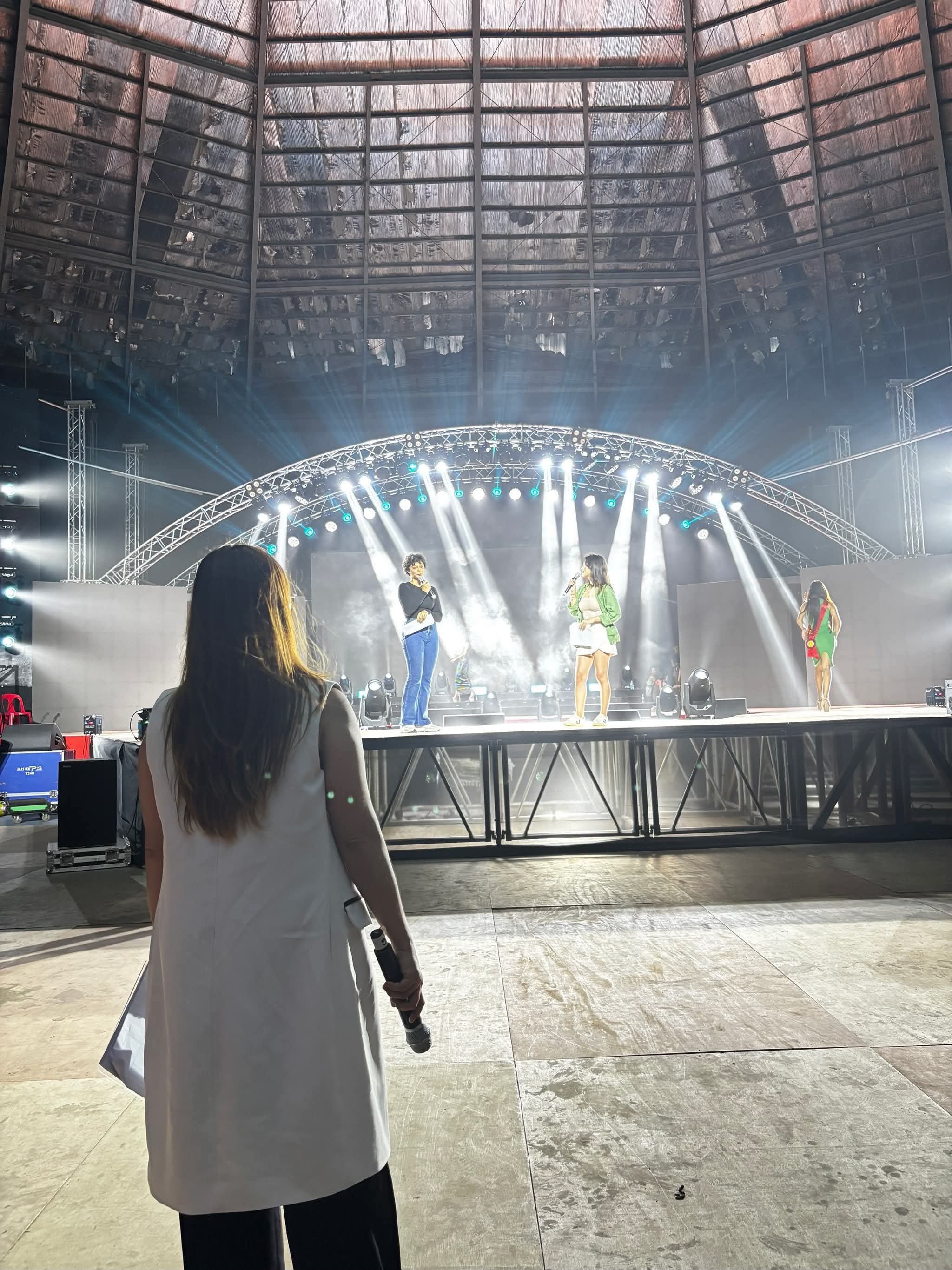 A coordinator watching a rehearsal from the floor of a covered arena, microphone in hand, as three performers run their spot under the lights