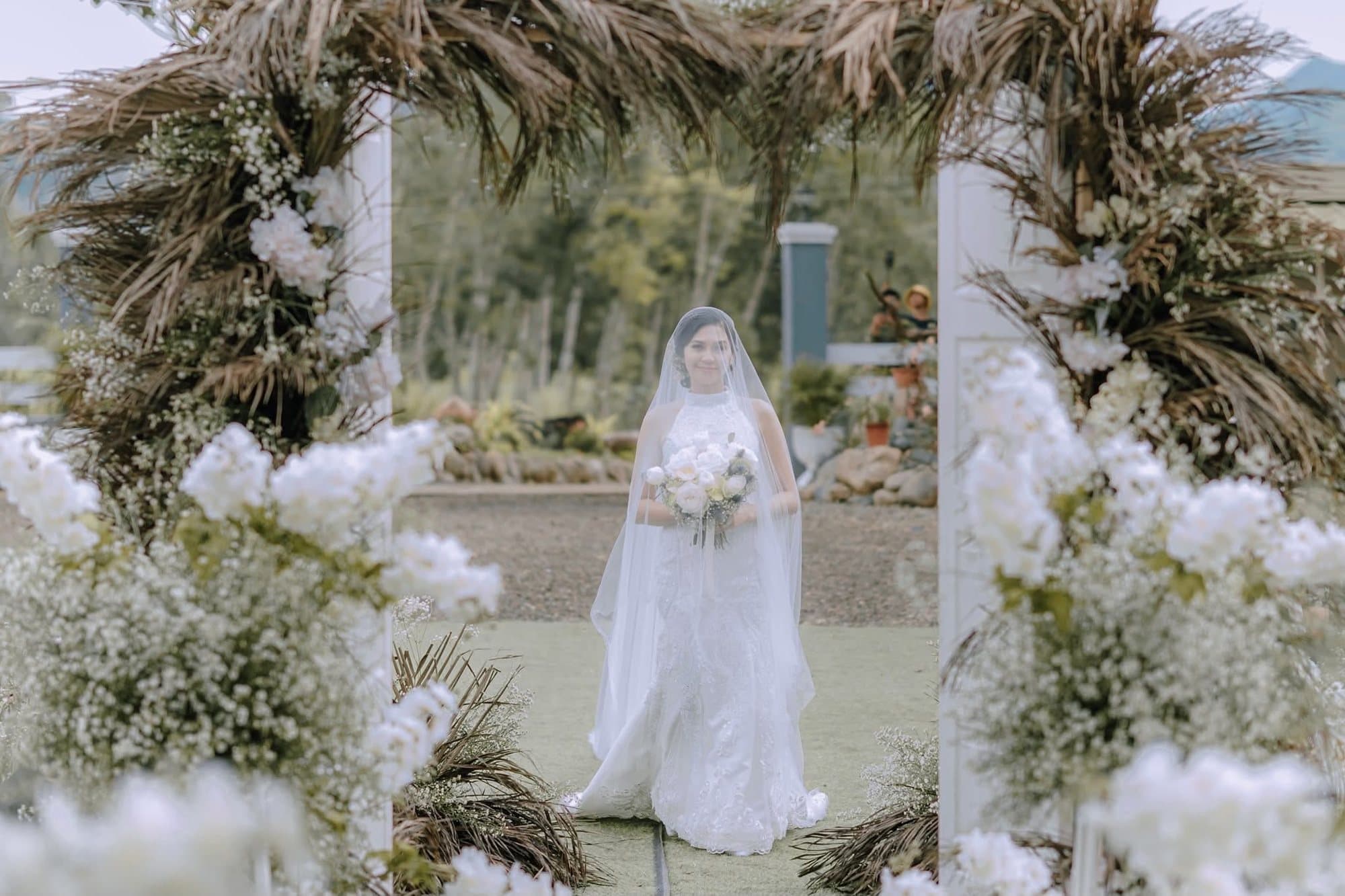 A veiled bride framed by a floral arch at an outdoor ceremony