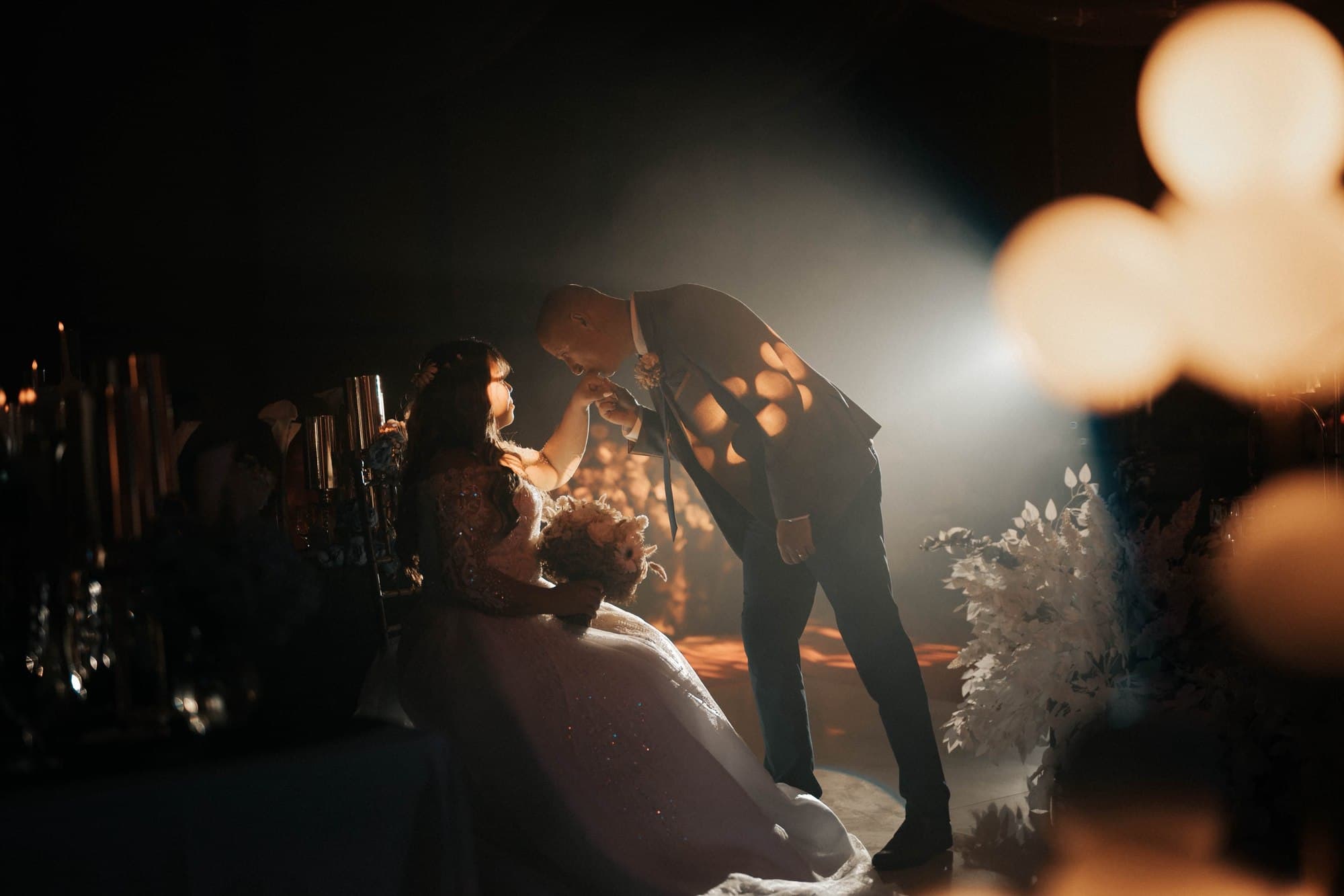 A groom bending to kiss his bride's hand at the head table, in candlelight